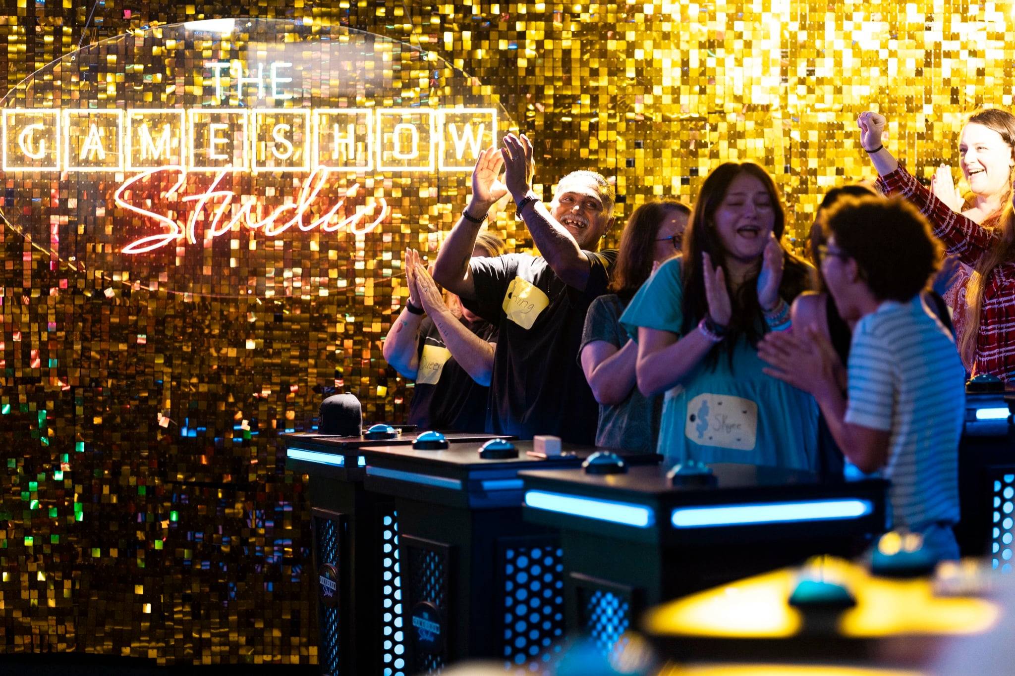 Adults and teens stand behind illuminated blue buzzers on a game show stage, cheering and laughing with raised hands.